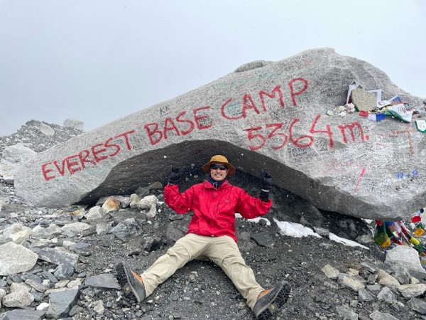 Man in climbing gear sitting in front of a rock with the words Everest Base Camp 5364 meters written on it.