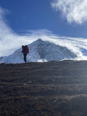 Man in climbing gear standing on a mountain with a snowy mountain peak in the background.