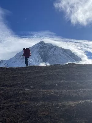 Man in climbing gear standing on a mountain with a snowy mountain peak in the background.